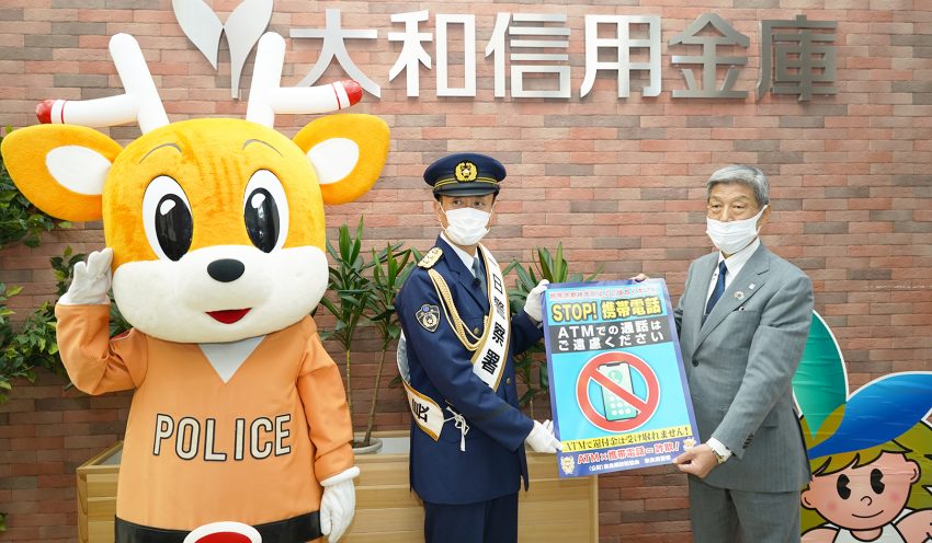 桜井警察署の一日警察署長岩本さん（中央）から特殊詐欺啓発ポスターを受け取る森川理事長（右）、左は奈良県警マスコットキャラクター「ナポくん」