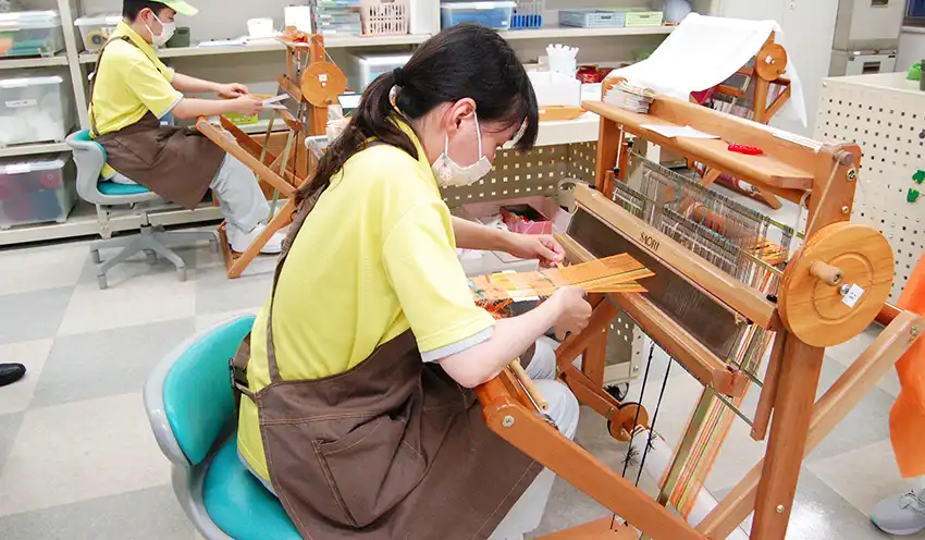 手織り機で織物を製作する高橋歩希さんら(伊予銀行事務センター、6月27日)