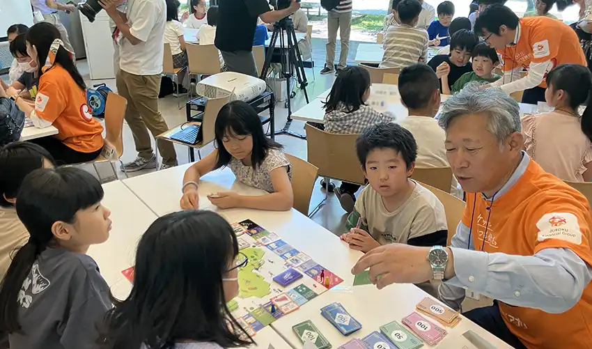 ボードゲーム「のぶながマネースタディ」を遊ぶ子どもと案内役の十六銀行員（８月３日、ぎふメディアコスモス）