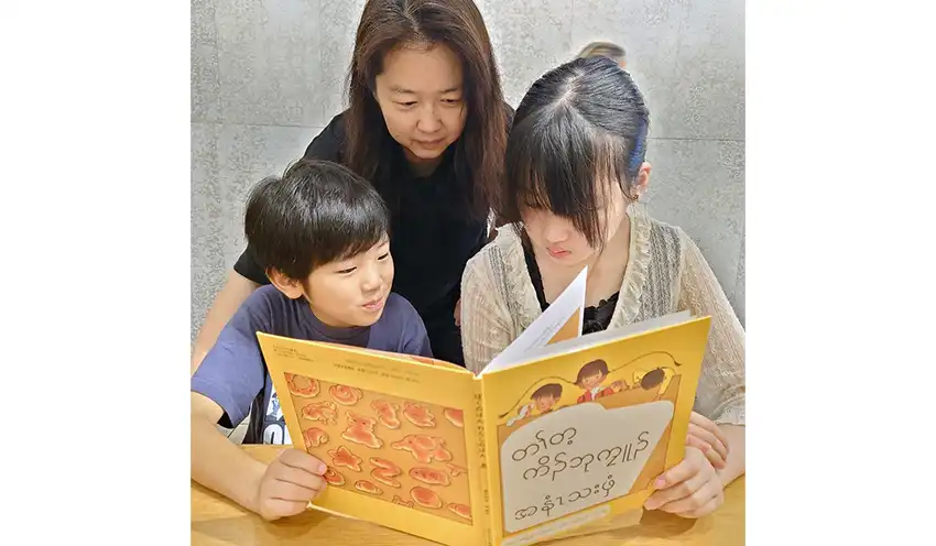 福音館書店の「ぼくのぱんわたしのぱん」にカレン語の翻訳シールを貼った久保さん（中央）と子ども2人（8月24日、久保さん宅、住友生命提供）