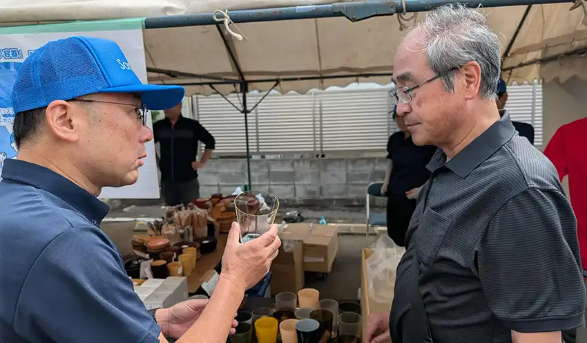 三義漆器店の曽根佳弘社長（左）からPLAグラスの説明を聞く菊地理事長（9月20日、会津商工信組本店駐車場、会津商工信組提供）