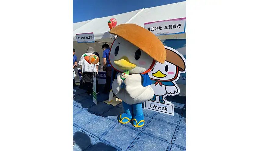 ブース前でポーズをとるしがの助。手にしているのは2026年に青森県で開催される大会のマスコット（滋賀銀提供）