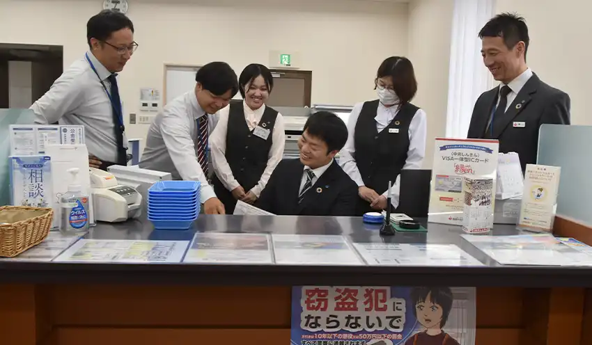 新入職員（左から４人目）に先輩職員らが教える様子を見守る檜山支店長（右、10月８日、島根中央信金松江北支店）